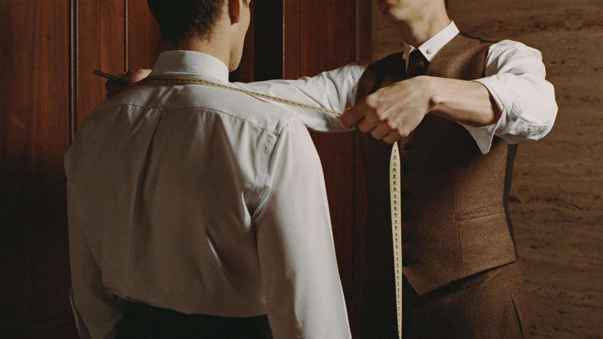A tailor measuring across a client's shoulders with a tape measure, against walnut panelling