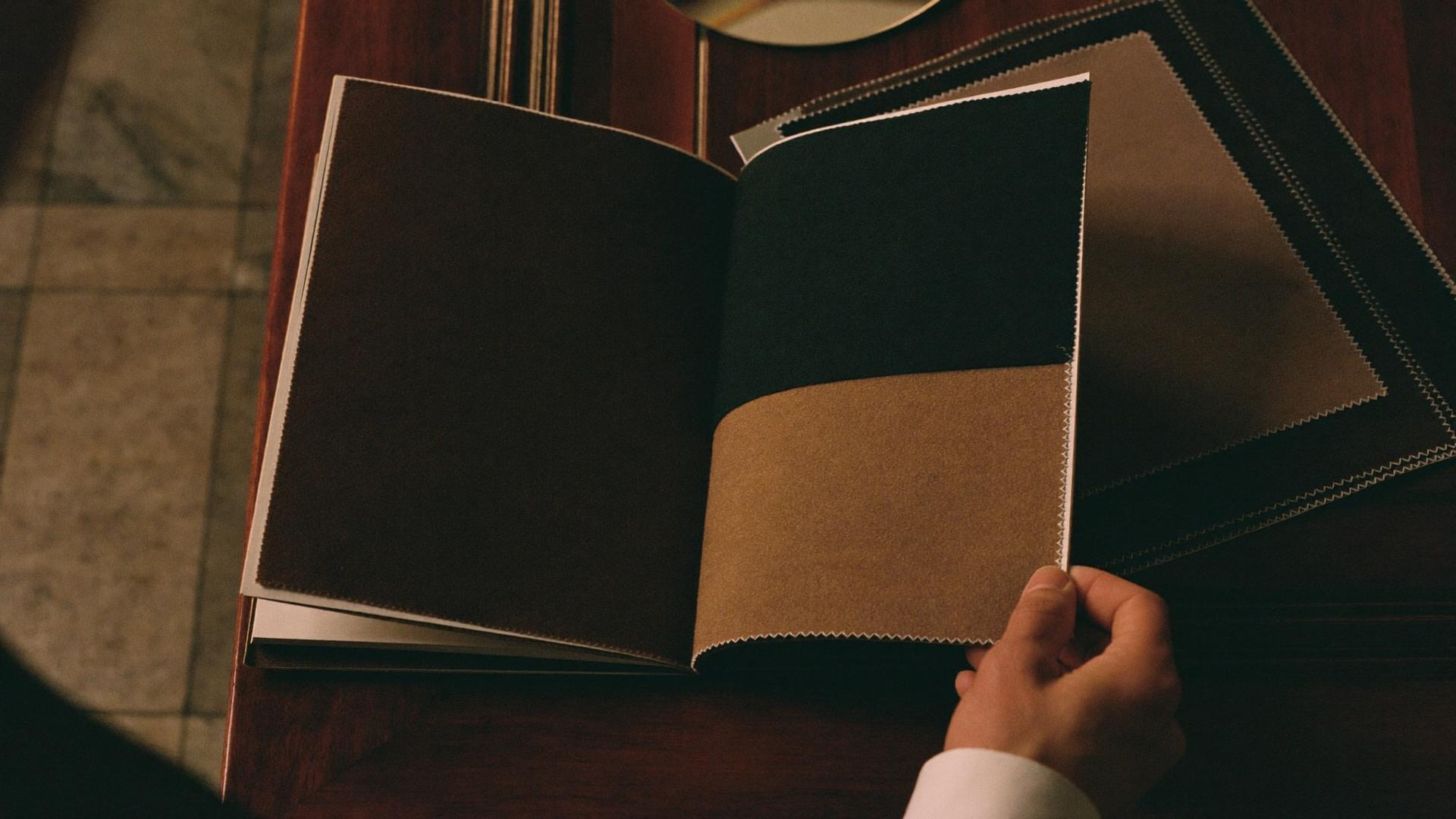 A hand turning the pages of a fabric swatch book on a walnut table