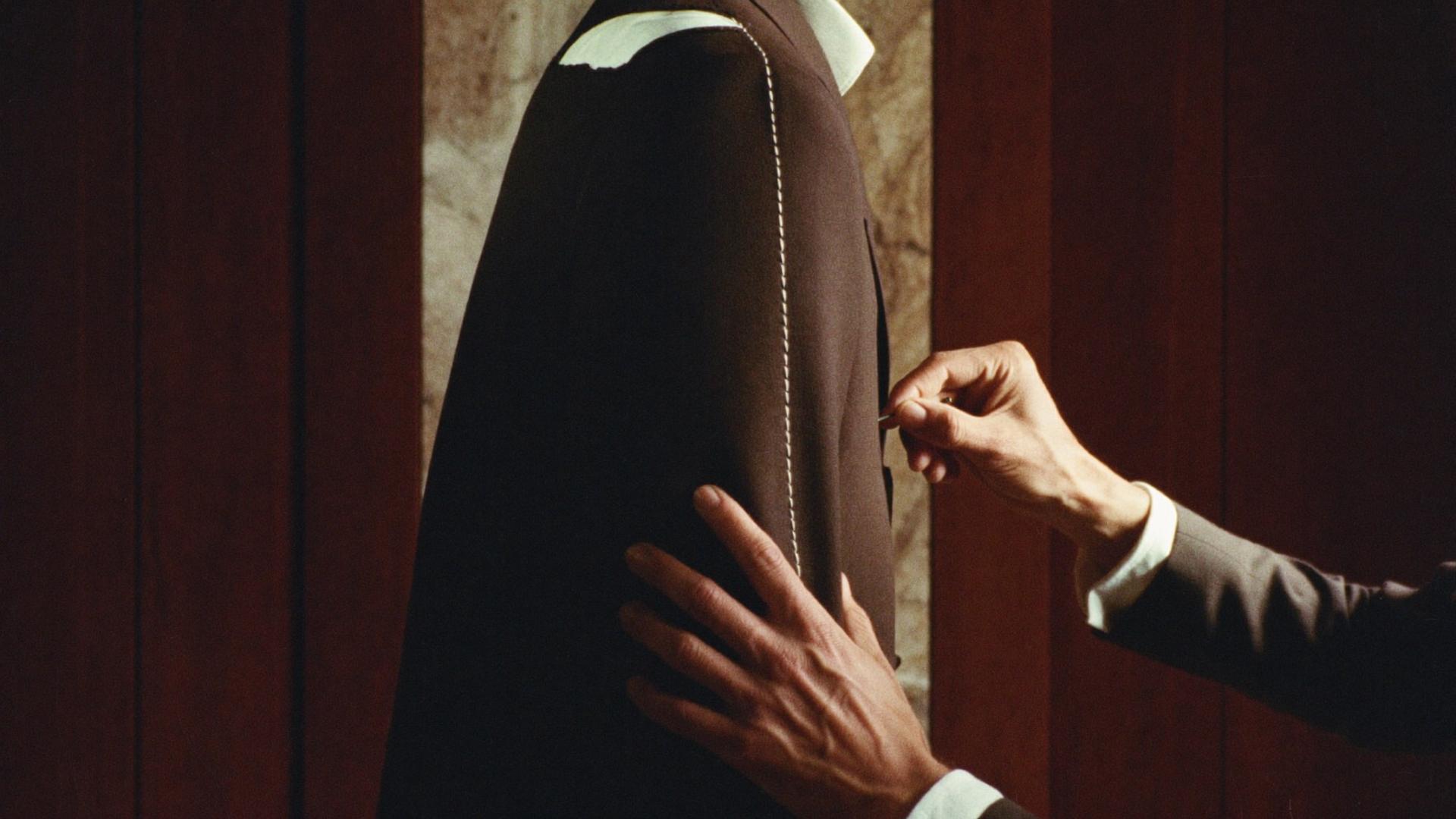 A tailor's hands adjusting a basted jacket at a fitting, white tacking along the shoulder