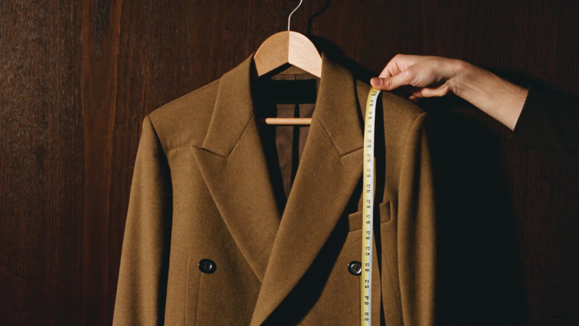 A finished double-breasted jacket on a wooden hanger, a tailor’s hand settling the lapel